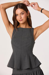 A woman is wearing a sleeveless, fitted, gray dress with a subtle checkered pattern. The dress features a round neckline and princess seams that create a tailored fit. It has a flared, ruffled peplum at the waist that extends to a pleated skirt with a ruffled hem. She accessorizes with large gold hoop earrings and a black bracelet on her right wrist. The woman has shoulder-length, wavy brown hair and is posing with her arms raised above her head against a plain, light background.